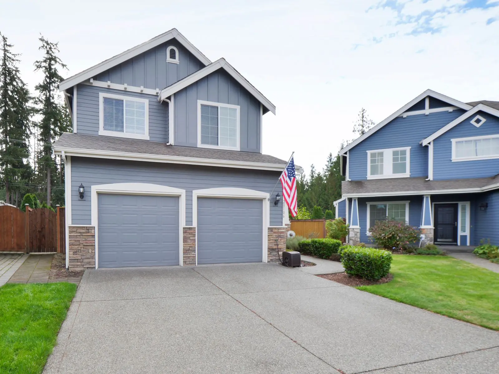 Modern blue house with double garage, American flag, landscaped yard, and neighboring homes in a suburban setting.